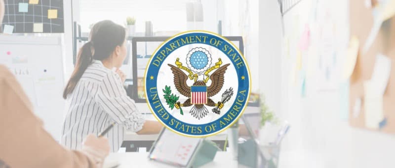 View of a woman working at a desk looking at a large monitor with lots of post-it notes on the wall. There is a white overlay with the U.S. Department of State seal in the middle.